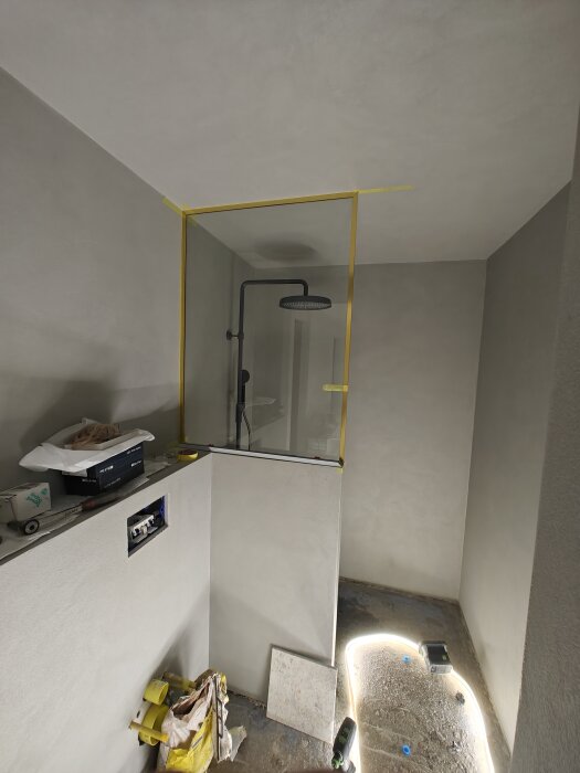 Renoveringsarbete pågår, badrum med duschvägg, byggmaterial syns, arbetslampor, ofärdigt, gult maskeringstejp, grått kakel.