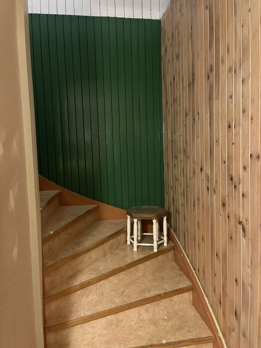 En trappa med träpanel, grön vägg, och en pall står på ett trappsteg, inomhus.