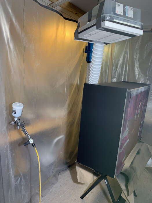 Verkstadsutrymme med ventilationssystem, målarpistol, upplyst vägg, plastskynken och svartmålad föremål på ställning.
