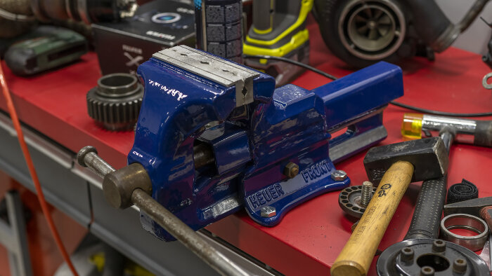 Blå skruvstycke på röd arbetsbänk med verktyg och mekaniska delar.
