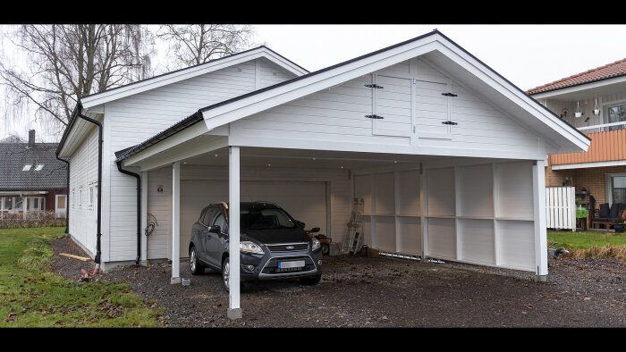 Vit carport med ett mörkgrått fordon, anslutet till ett hus, gråmulen himmel, bostadsområde i bakgrunden.