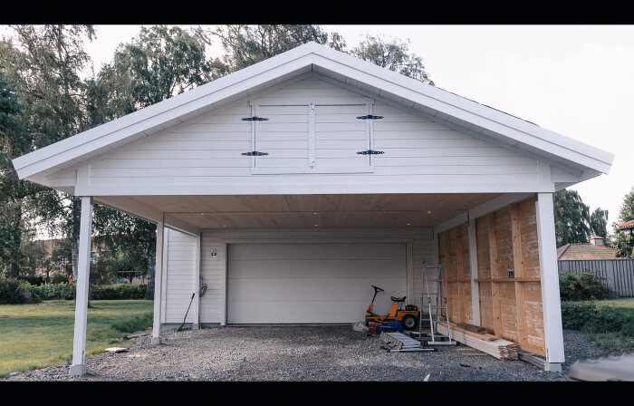 Vit carport med oöppnad garageport, verktyg, byggmaterial, gräsklippare, stege, under konstruktion, trädgård i bakgrunden.