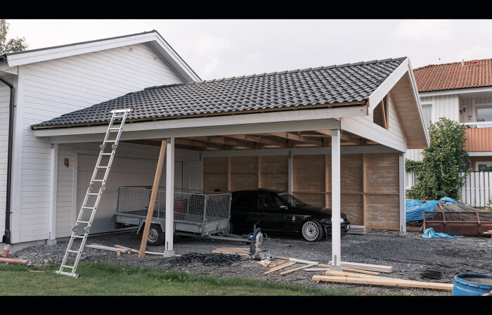 Garage under konstruktion, bil inuti, stege, släpvagn, byggmaterial utspridda, bostadsområde, grå molnig himmel.