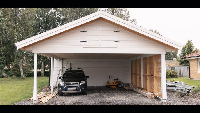 Tvåbilsgarage i trä med bil, gräsklippare, ved och byggmaterial under öppen del.