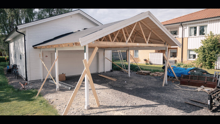 Nybyggd carport med trästomme vid vit husfasad, byggmaterial runt, stöttor, halvtak, byggprojekt i trädgård.