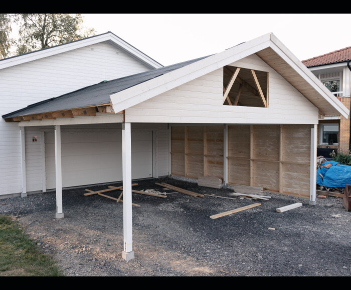 Ett ofärdigt carportbygge med vita plankor, byggmaterial på marken, i villaområde.