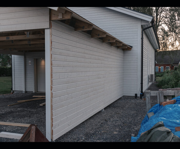 Nybyggd carport vid hus, byggmaterial, träbjälkar, vit fasad, skymning, grusgolv, plåtarbete, trädgård synlig bakom.