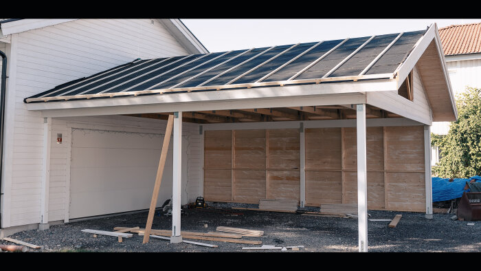 Nybyggd carport med svart tak nära vit husvägg, byggmaterial på marken, lövträd.