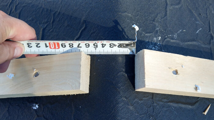 Hand håller måttband över träbitar med spikhål, mäter avstånd, byggprojekt, blå textilbakgrund.