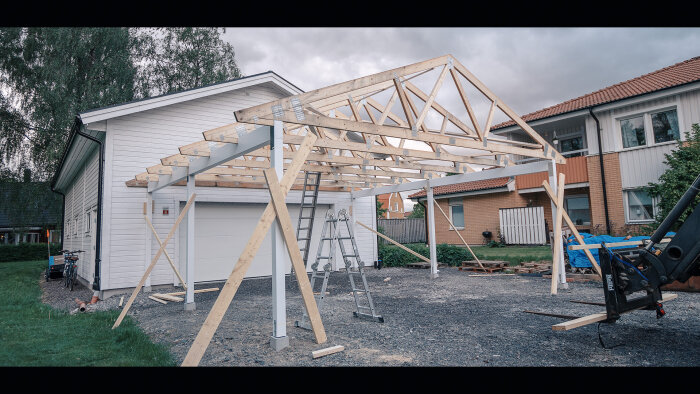 Byggnation av trätakstomme vid vit huskropp, grå grund, med stege och byggmaskineri i förgrunden.