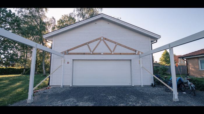 Vit garagebyggnad, grå garagedörr, träkonstruktion synlig, cyklar parkerade. Trädgård och andra hus i bakgrunden.