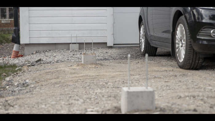 Delvis parkerad bil, grusunderlag, konstruktion med betongplintar och armeringsjärn framför garage.