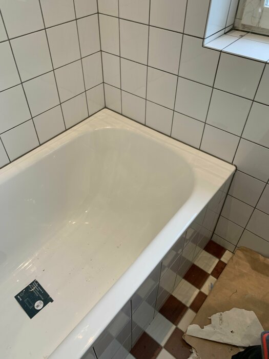 Vitt badkar, vita kakelväggar, brun mosaikgolv, renoveringsarbete, smuts, byggmaterial, spegelkant synlig.