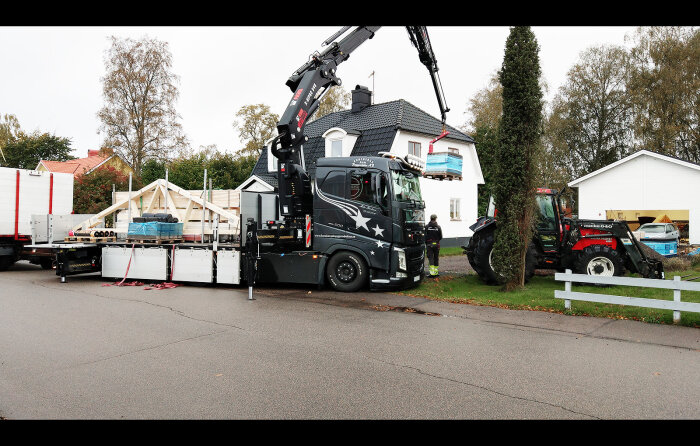 Lastbil med kran lyfter byggmaterial vid husbygge, arbetare i bakgrunden, bostadsområde.