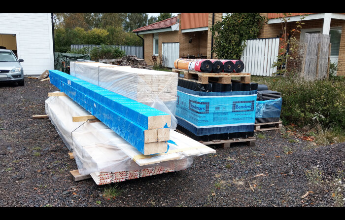 Byggmaterial på pallar, isolering och takpannor, utomhus vid byggarbetsplats eller renovering, bil till vänster.