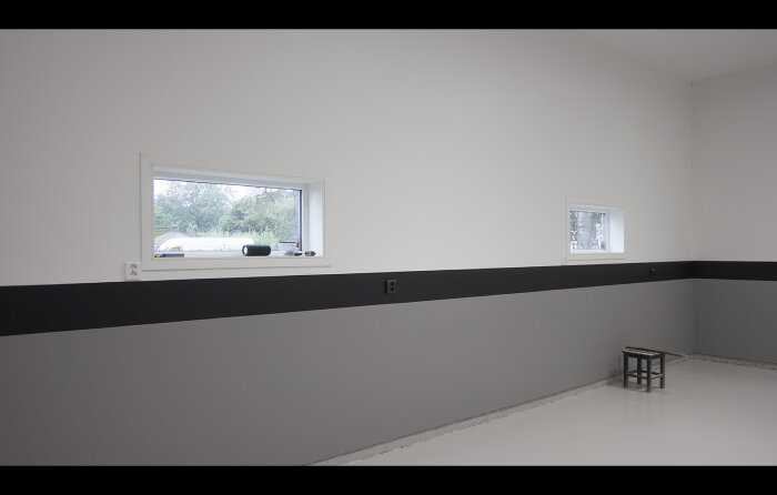 Minimalistiskt rum, vita väggar med svart bård, två fönster, en pall, blankt golv, neutralt ljusinsläpp.