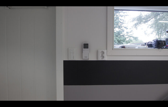 Vägg med fönster, termostat, strömbrytare, uttag, svart rand, del av kamera utomhus.