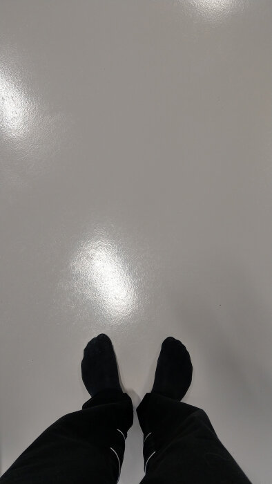 Person står på blankt golv i svarta strumpor och byxor, perspektiv uppifrån och ner.