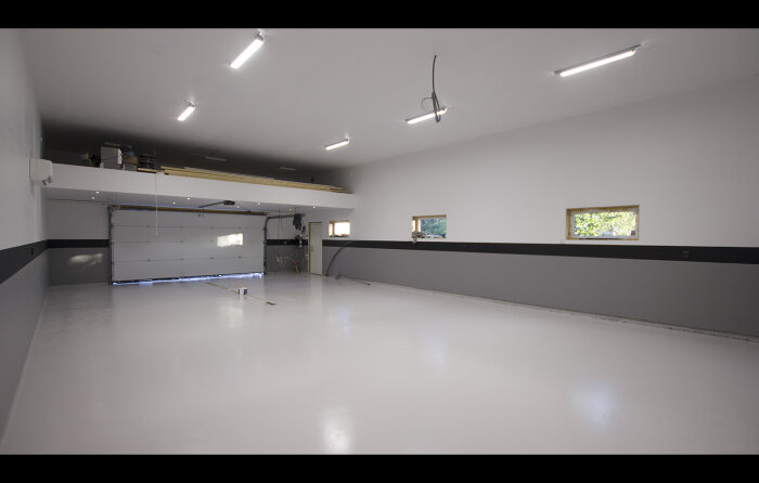 Stor, tom lokal med vit blank golvbeläggning, garageport, fönster och takbelysning.