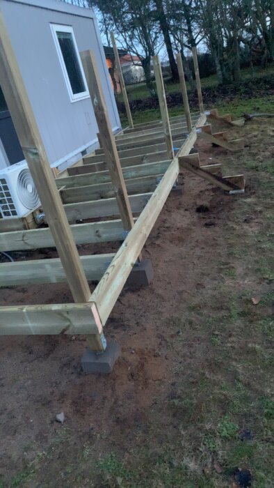 Träkonstruktion för däck eller altan under uppbyggnad vid hus, med stolpar och bjälkar på stödblock.