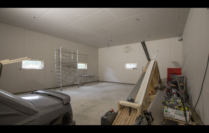 Inomhus, garage eller verkstad under ombyggnad, verktyg och material synliga, delvis parkerad bil, stegar, oslipat golv.