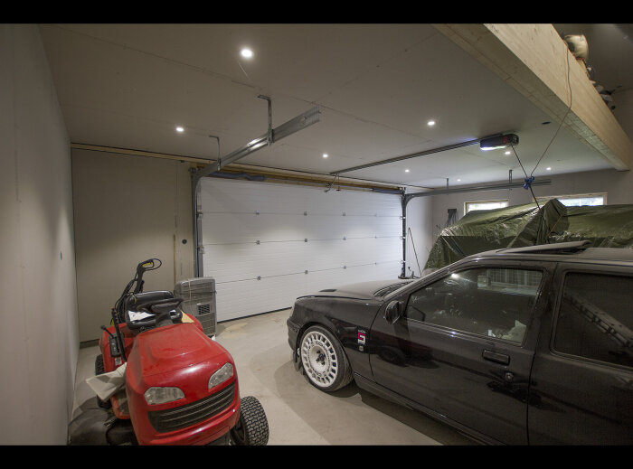 Ett garage med två bilar och en gräsklippare, upplyst av taklampor, med stängd port.