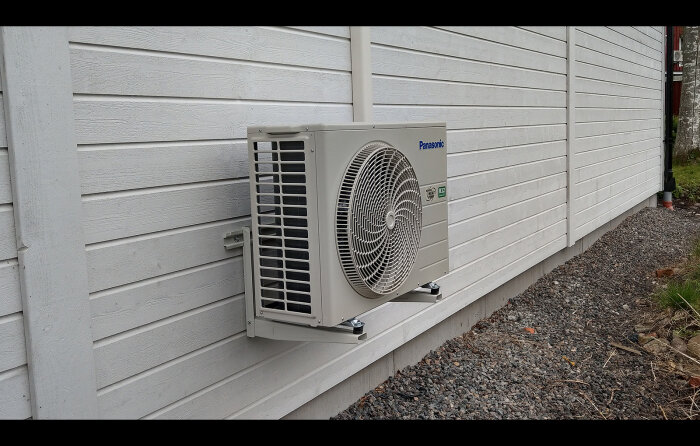 Luftvärmepump monterad på vit trävägg utomhus bredvid grus och grönska.