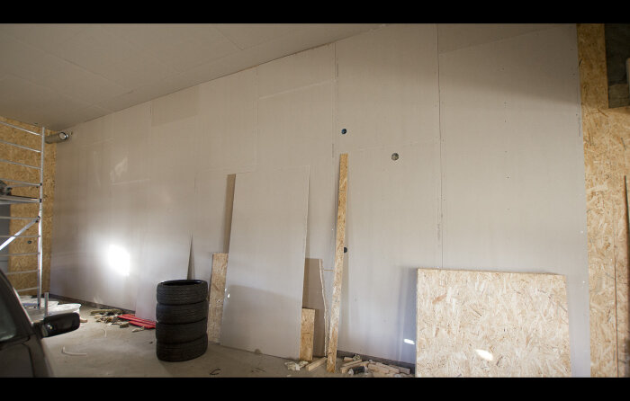 Inomhusbyggarbetsplats med gipsskivor, OSB-paneler, däckstapel och byggmaterial. Oavslutad konstruktion och inredning.
