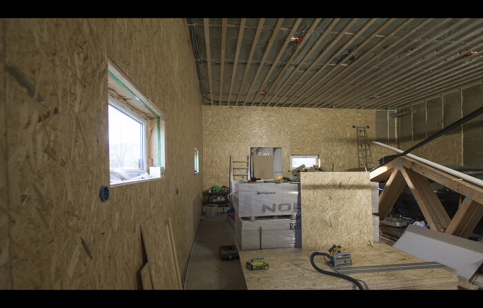 Byggarbetsplats inomhus med OSB-väggar, byggmaterial, verktyg och oisolerad takkonstruktion.