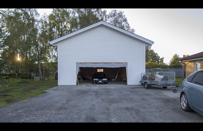 Vit byggnad med öppen garageport, bil och släpvagn framför, träd och buskar i skymningen.