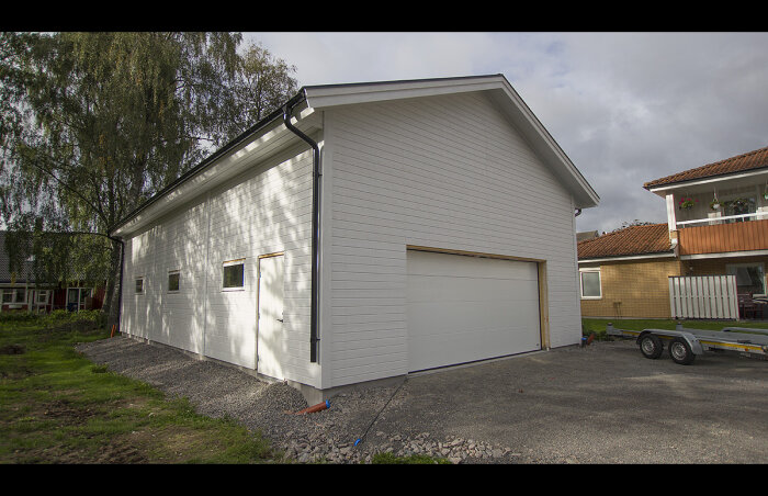 Vit garagebyggnad med två fönster, en stor port, grus och släpvagn framför.