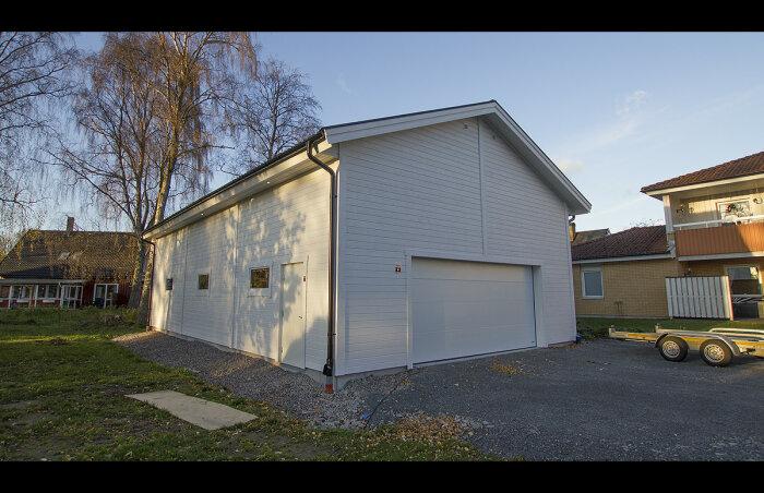 Vit garagebyggnad med dubbelport, hus i bakgrunden, släpvagn, grus, gräs, träd.