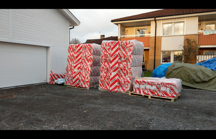 Paket inslagna i varningsband ligger utomhus nära ett bostadshus och en garagebyggnad. Gråmulet väder.