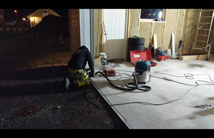 Person arbetar utomhus nattetid med elverktyg, kablar och byggmaterial på en garageuppfart.