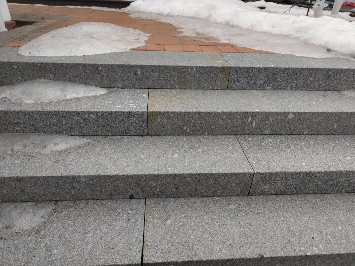 Trappsteg av grå sten utomhus med snöfläckar och bakgrund av smältande snö.