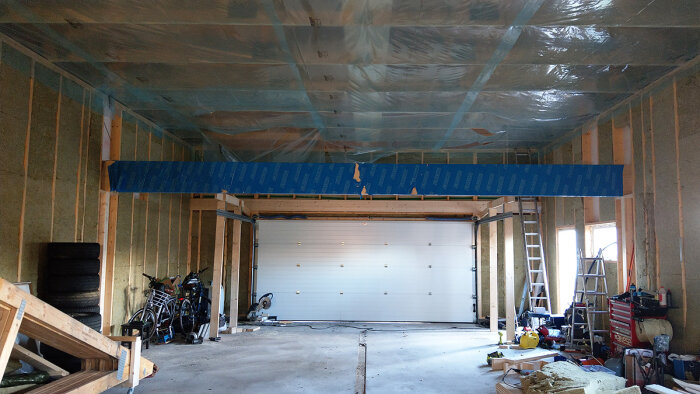 Garage under renovering, isolering synlig, garageport stängd, byggmaterial och verktyg spridda, stegar, plasttäckt tak.