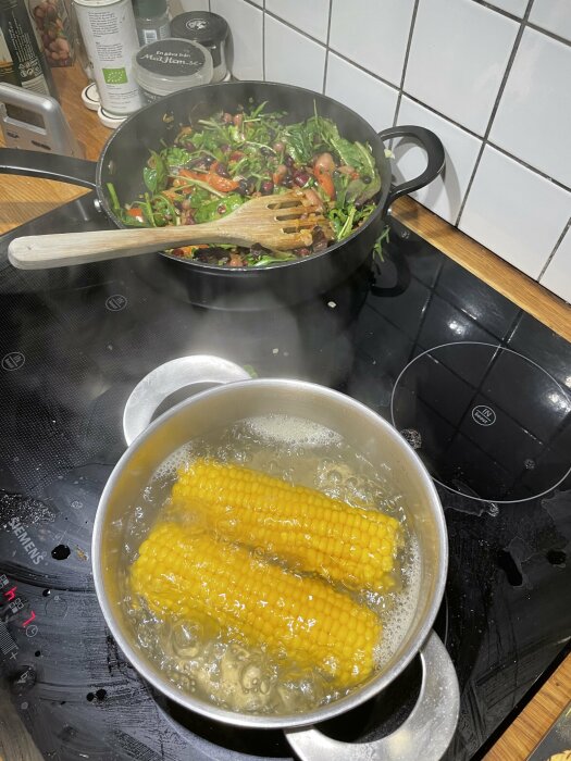 Kokande majs i kastrull, sallad i wokpanna på spishäll, matlagning, ångande, köksmiljö.