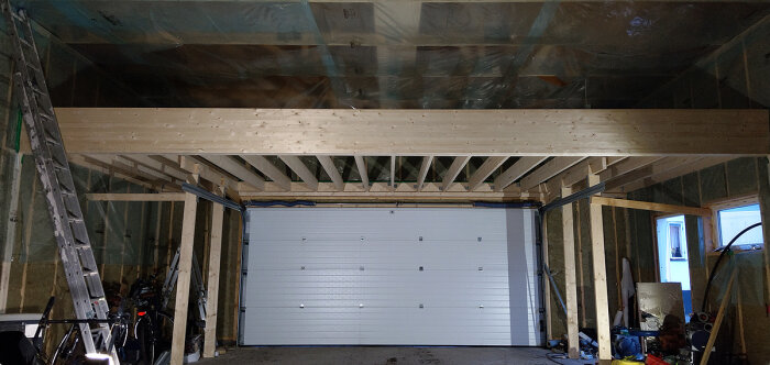 Nybyggd garageinteriör med isolering, träbjälkar och en stängd garageport. Stege och föremål förvaras inuti.