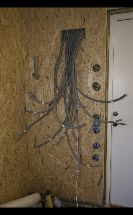 Installation under arbete: kablar och rör på spånskiva, genomföringshål, ouppackade delar, dörr till höger.