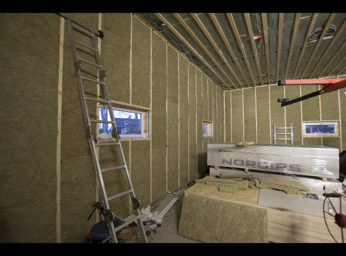 Byggarbetsplats inomhus med isolering, stegar, trästomme och material märkt "NORGIPS". Oavslutat konstruktionsprojekt.
