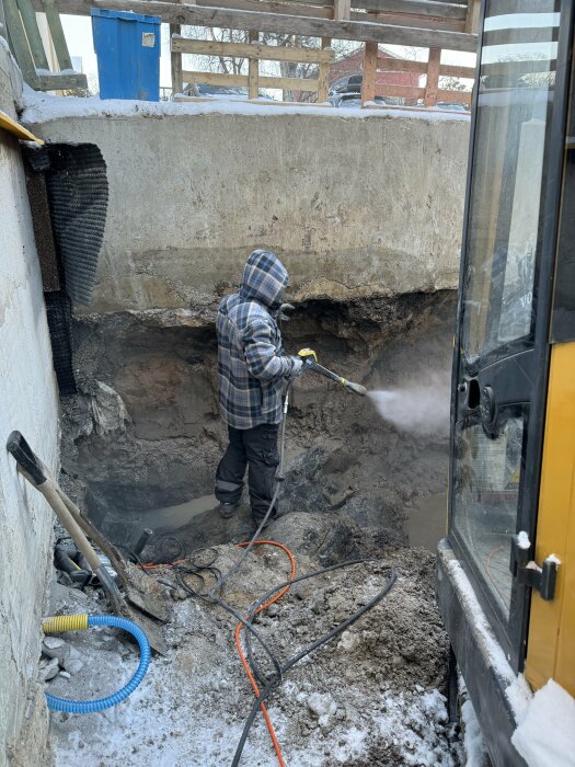 Person i huvjacka använder högtryckstvätt i grävd grop vid husgrund, vinter, verktyg, rör, snö.