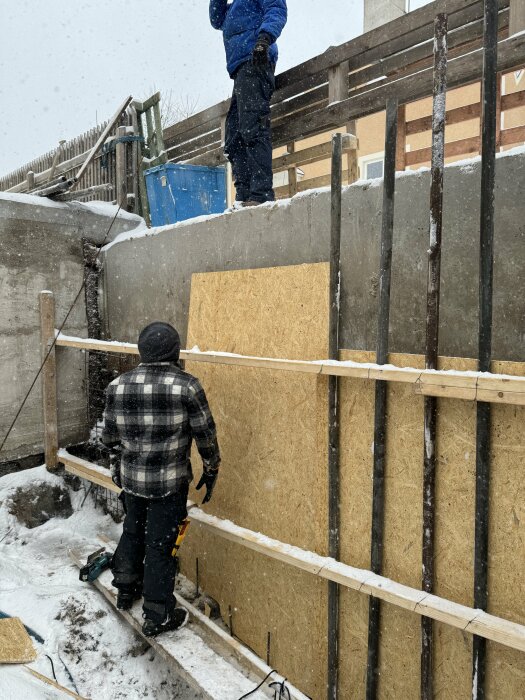 Två personer arbetar utomhus med isolering och byggnation under snöfall.
