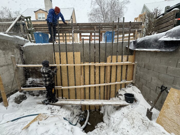 Två arbetare bygger en träform i snöväder för betonggjutning vid en byggarbetsplats.