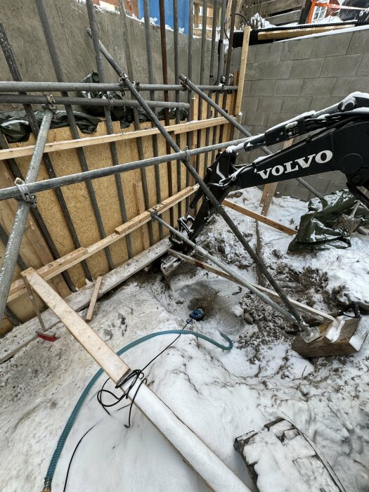 Byggarbetsplats med snö, en Volvo grävmaskinarm, träplankor, stängsel och byggmaterial på marken.