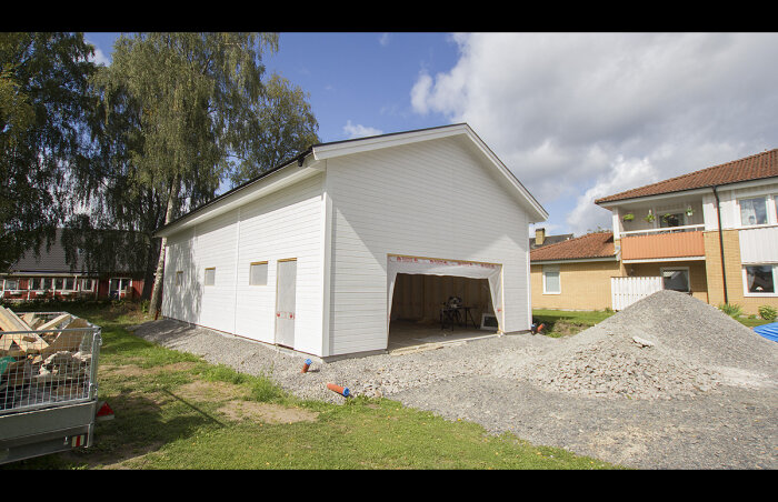 Vit fristående byggnad, öppen garageport, byggmaterial, cykel, grus, bostadshus bakgrund, delvis molnig himmel.