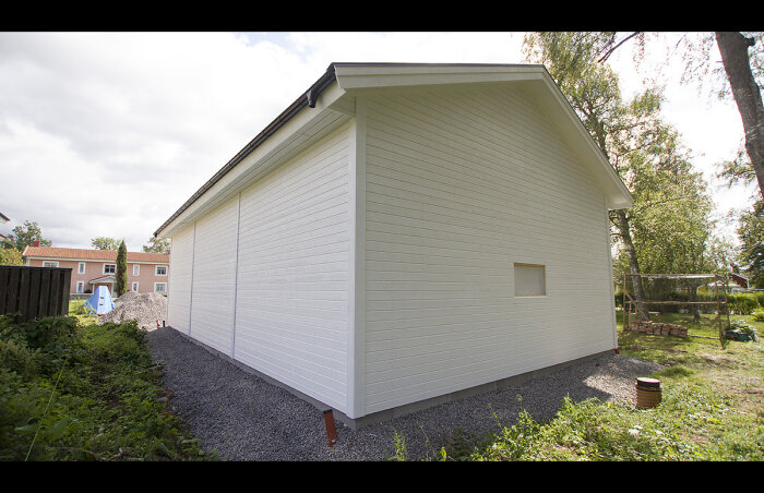 Litet vit hus med lutande tak, grus runtom, trädgård och blå himmel i bakgrunden.