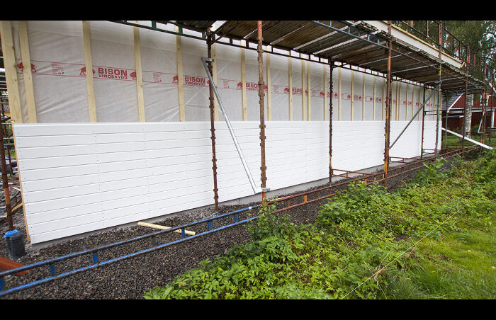 Byggnadsställning vid hus under renovering, isoleringsmaterial synligt, omgiven av grönska, halvfärdig fasad.