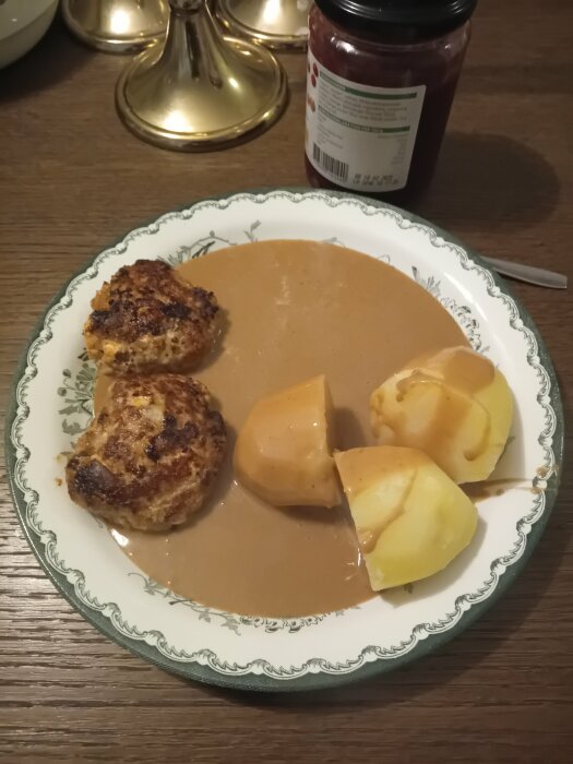 En tallrik med köttbullar, potatis och brunsås, en burk lingonsylt och ett ljusstake i bakgrunden.