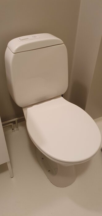 En vit toalettstol med öppet lock i ett badrum med beige väggar.