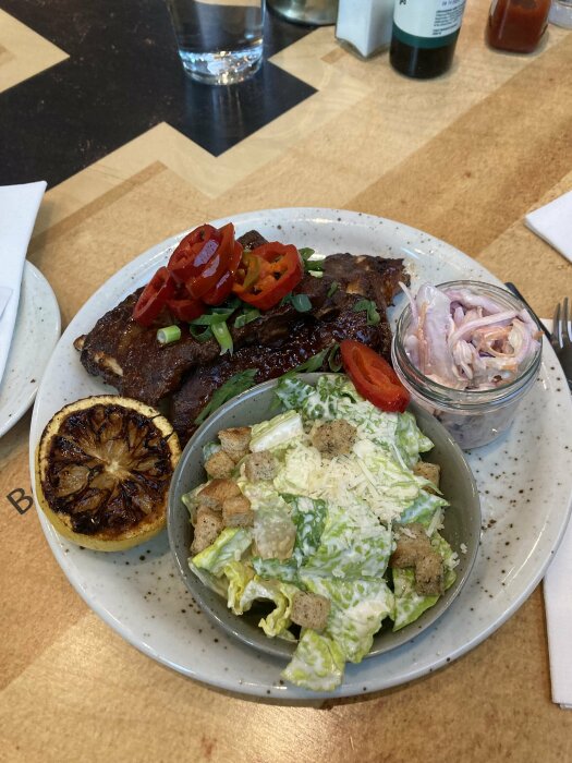 Måltid med revbensspjäll, Caesarsallad, picklad lök och grillad citron. Glas vatten. Trämönstrat bord.